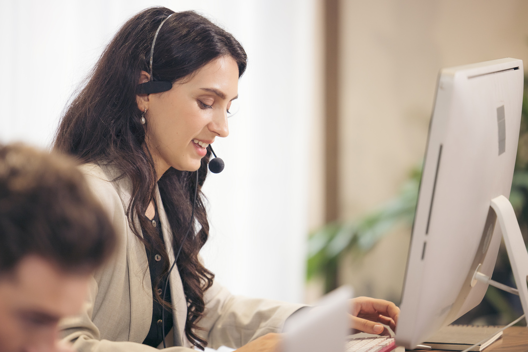 Na imagem tem uma mulher realizando tipos de atendimento ao cliente, onde ela está com um fone com microfone de frente para um computador.